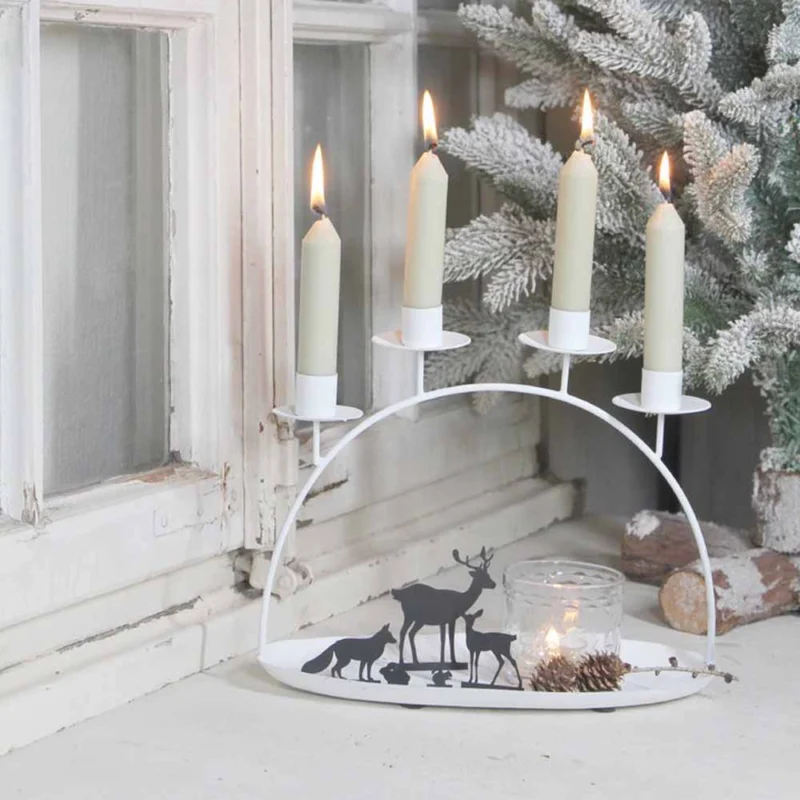 Die VL Home Schale mit Kerzenbogen für vier Stabkerzen ist beleuchtet und zeigt Tiersilhouetten auf einem weißen bogenförmigen Halter, der neben einem Milchglasfenster und einem schneebedeckten künstlichen Baum platziert ist.