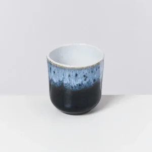 Blau und schwarz glasierte Keramiktasse vor einem hellgrauen Hintergrund.