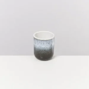 Keramiktasse mit einem Farbverlauf von Schwarz zu Weiß auf einem weißen Tisch vor grauem Hintergrund.