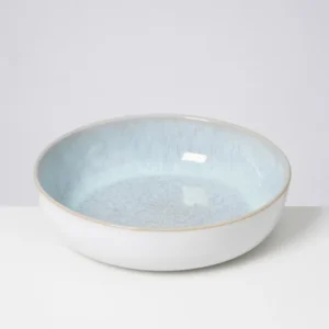 Eine flache, runde Schüssel mit blauer Innenseite und weißem Rand vor grauem Hintergrund.