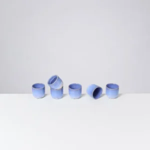 Sechs blaue Keramiktassen stehen und liegen auf einem weißen Tisch vor einem grauen Hintergrund.