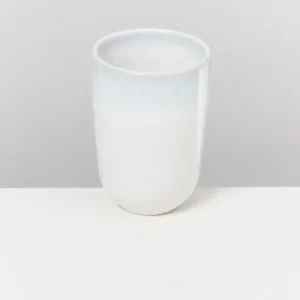 Weißer Keramikbecher mit blauer Oberkante auf einem weißen Tisch vor grauem Hintergrund.