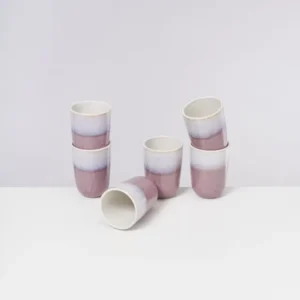 Sechs lila-weiße Keramikbecher, fünf aufrecht und einer liegend, auf einem weißen Tisch vor einem grauen Hintergrund.