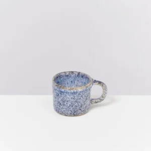 Blaue, gesprenkelte Keramiktasse mit Henkel auf einem weißen Tisch vor grauem Hintergrund.