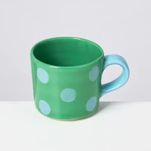Grüne Tasse mit blauen Punkten und blauem Henkel vor einem weißen Hintergrund.