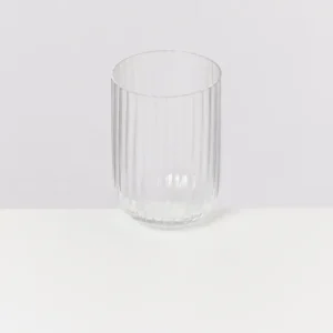 Ein geripptes, klares Glas steht auf einer weißen Oberfläche vor einem grauen Hintergrund.