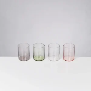Vier transparente, farbige Gläser in Lila, Grün, Klar und Rosa auf einem weißen Tisch vor grauem Hintergrund.