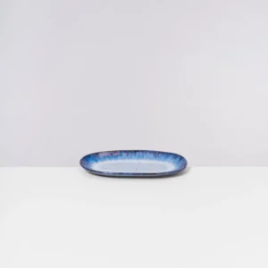 Eine ovale, blau-weiße Keramikplatte auf einem weißen Tisch vor grauem Hintergrund.