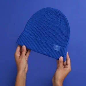 Hände halten eine blaue Strickmütze vor einem blauen Hintergrund.